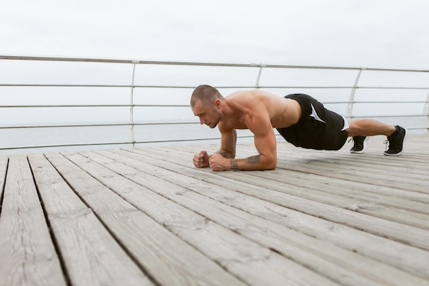 ビーチで板の運動をしている裸の胴体を持つハンサムな筋肉質の男