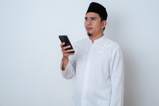 Foto bell'uomo musulmano che indossa abiti musulmani e che tiene il suo telefono che guarda lontano contro il muro bianco