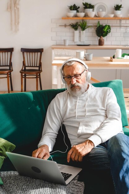 Handsome old man dressed in hoodie and eyeglasses is using laptop and smiling while listen music sit