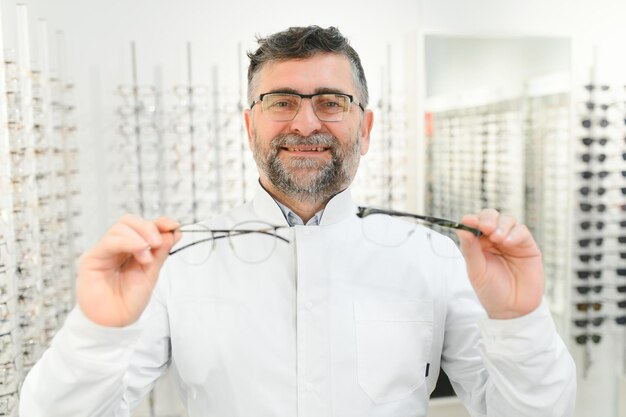 시험을 위해 안경을 들고 있는 잘생긴 안과의사 검안사가 손과 안경에 초점을 맞춘 작은 심도의 안경 이미지를 착용하겠다고 제안합니다.
