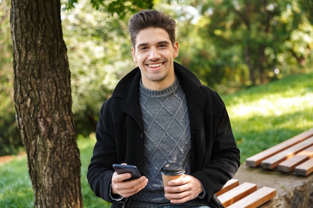 사진 휴대 전화 마시는 커피를 사용 하여 녹색 공원에서 야외 산책 캐주얼 의류에 잘 생긴 낙관적 젊은 행복 한 사람.