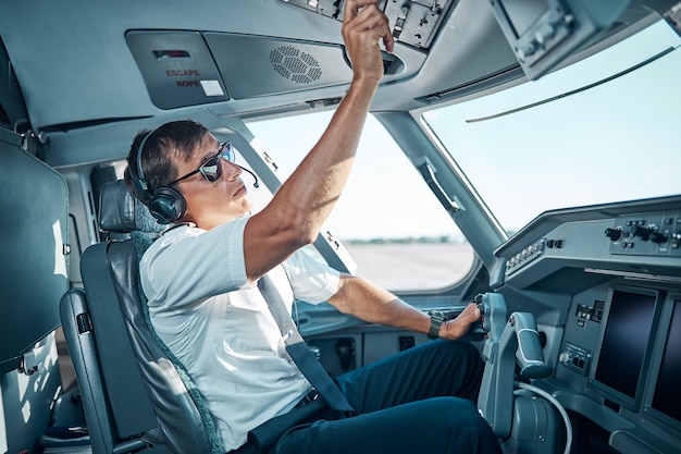 Handsome pilot in glasses and earphones is sitting inside plane at control and preparing for departure