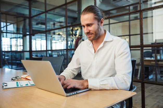 handsome pleased man with stubble working with laptop while sitting at table in modern office