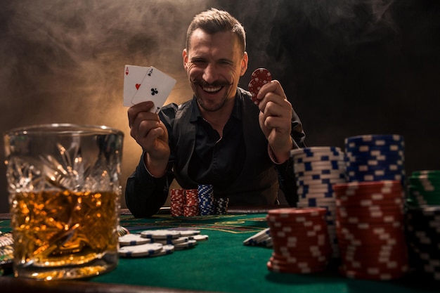 Handsome poker player with two aces in his hands and chips\
sitting at poker table in a dark room full of cigarette smoke.