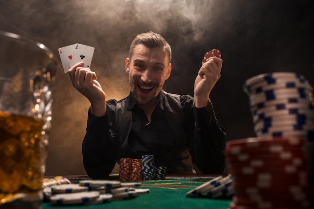 Handsome poker player with two aces in his hands and chips sitting at poker table in a dark room full of cigarette smoke.