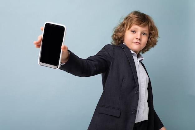 Bel ragazzo positivo con i capelli ricci che indossa tuta tenendo il telefono isolato su sfondo blu guardando la fotocamera e mostrando smartphone con schermo vuoto