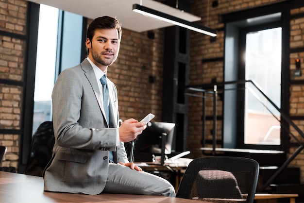 Handsome positive businessman sitting on office desk holding smartphone and looking at camera