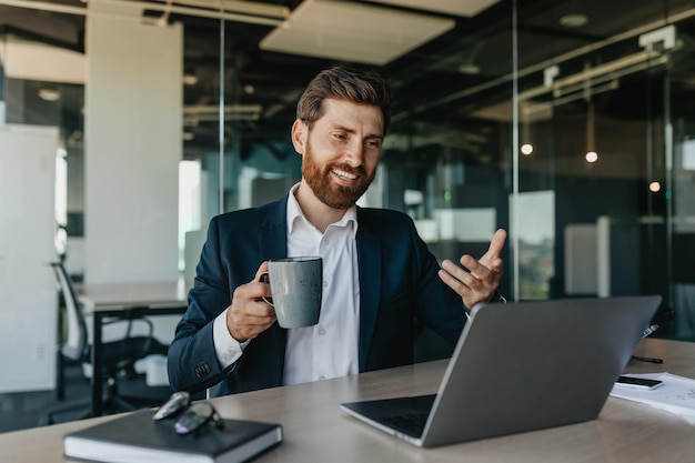 Handsome positive caucasian businessman in suit sitting in office talking with business partners via laptop