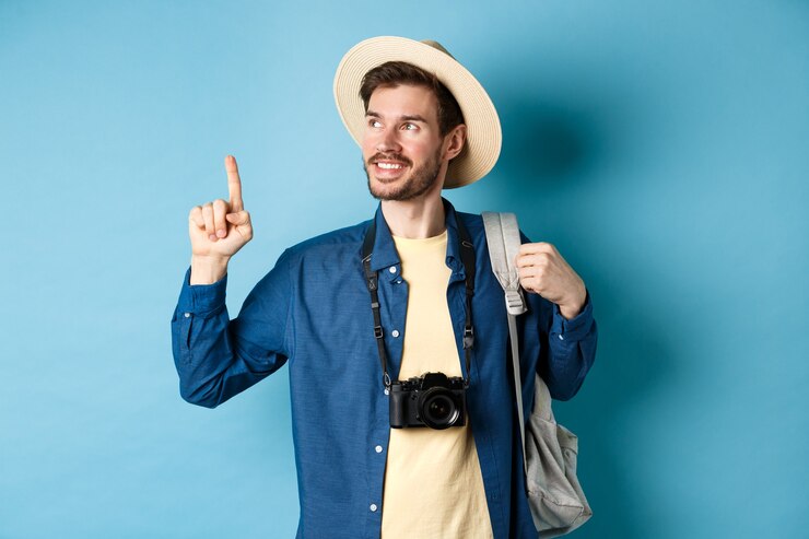 Photo handsome positive guy on vacation, wearing summer hat, looking and pointing up at logo, smiling pleased, standing with camera and backpack on blue background.