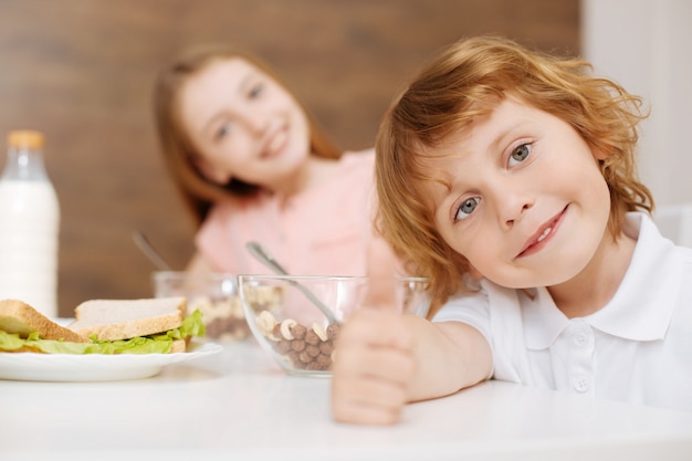 Handsome positive nice kid sitting at the table with his sibling and eating the first meal of the day and enjoying it