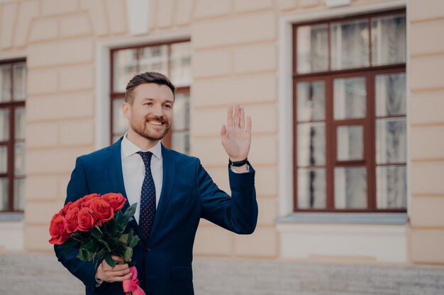 Handsome positive young man dressed in classic blue suit and tie with white shirt greeting his business partner outdoors, holding bouquet of red roses in hand while standing alone next to building