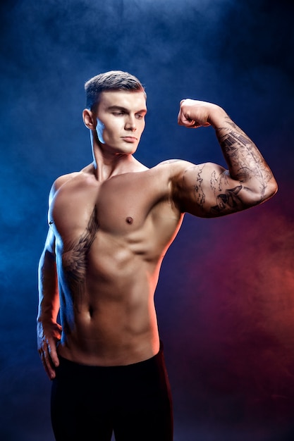 Handsome power athletic man bodybuilder. Fitness muscular body on dark smoke background. Perfect male. Awesome bodybuilder, tattoo, posing. 