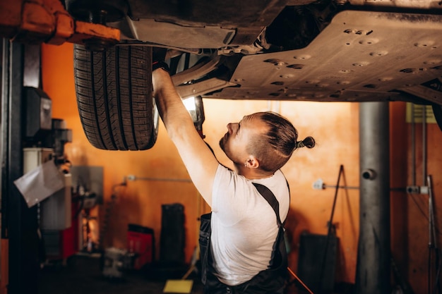 Handsome Professional Car Mechanic is Investigating Rust Under a Vehicle on a Lift in Service Repairman is Using a LED lamp and Walks Towards Specialist is Wearing Safety Glasses Modern Workshop