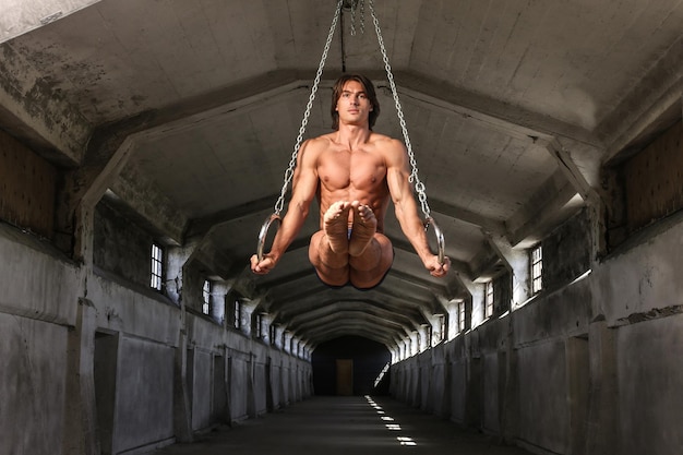 Un bel ginnasta professionista con un bel corpo muscoloso si allena su anelli da ginnastica nell'edificio industriale abbandonato, mostra la posizione statica