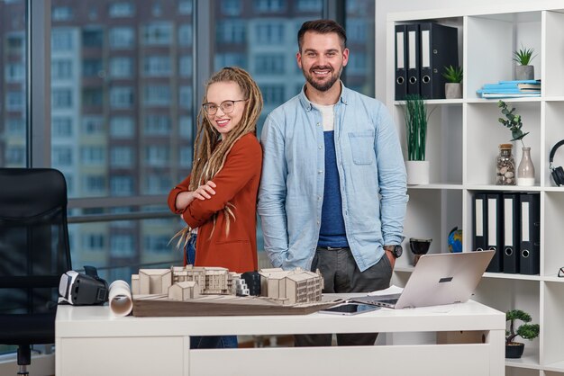 Handsome professional young engineer stands back to back with pretty female colleague with dreadlocks at design bureau.