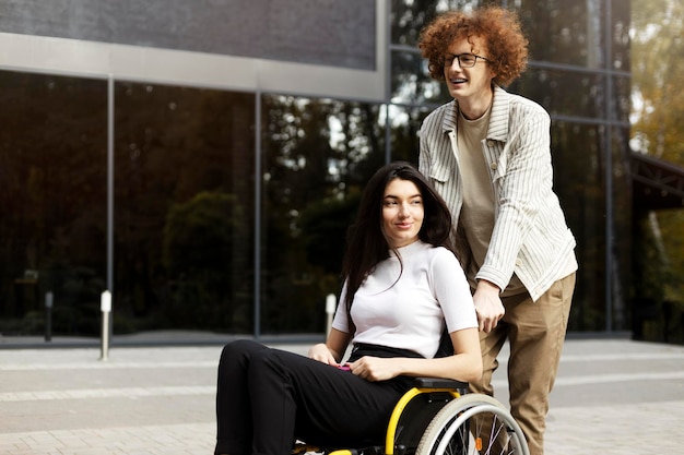 Handsome redhaired curly guy in glasses and braces carries a wheelchair with a girl A beautiful brunette sits and looks away on the street outdoors