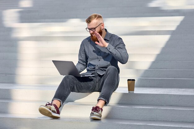 Handsome redhaired freelancer man with a beard sits on the street and works remotely using a laptop