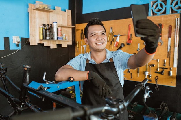 A handsome repairman smiling in an apron while taking a selfie using a cellphone while repairing a b...