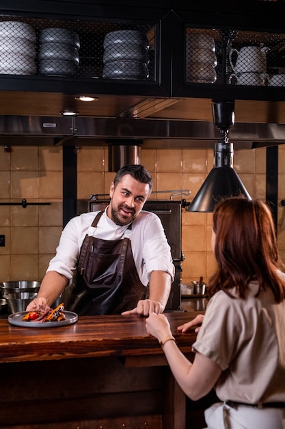 Handsome restaurant chef in apron standing at counter in open kitchen and telling about dish to guest