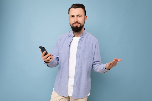 handsome sad young brunette unshaven man with beard wearing stylish white t-shirt and blue shirt isolated over blue background with empty space holding in hand and using phone messaging sms looking at