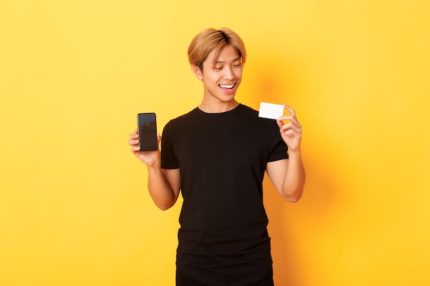 Handsome satisfied asian guy holding credit card and showing smartphone screen