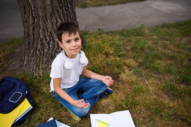 Handsome school child boy sitting in lotus position on the green grass of public park and meditating , looking up at the camera, cute smiling. Workbooks and school supplies lying on the grass