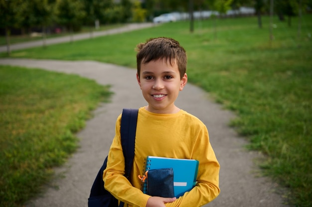 A handsome schoolboy with a school bag and notebooks stands on the path in the city park after school. Portrait of a cute adorable schoolboy