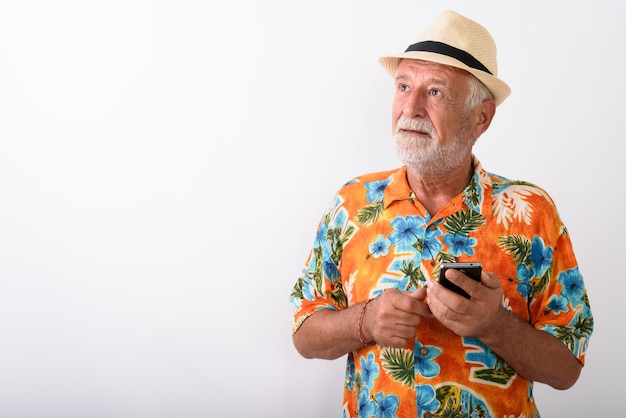 handsome senior bearded tourist man thinking while holding mobile phone and wearing hat on white