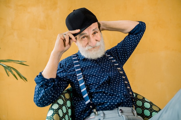 Uomo senior bello in camicia blu scuro, bretelle e cappuccio nero dei pantaloni a vita bassa che posano nello studio, sedentesi davanti alla parete gialla. uomo anziano alla moda alla moda su fondo giallo