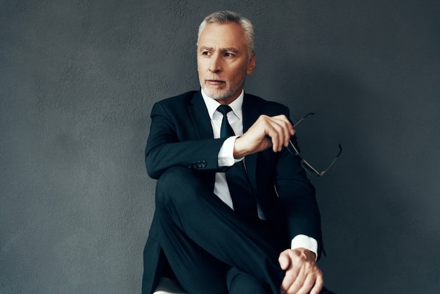 Handsome senior man in elegant full suit looking away while sitting against grey background