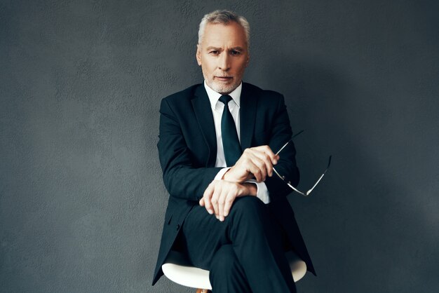 Photo handsome senior man in elegant full suit looking at camera while sitting against grey background