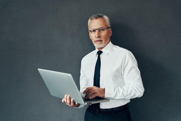 Handsome senior man in elegant shirt and tie using laptop and looking at camera while standing against grey background