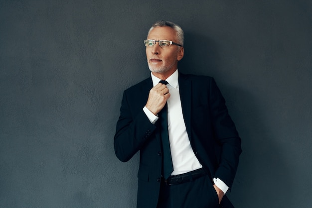 Handsome senior man in full suit looking away and adjusting tie while standing against grey background