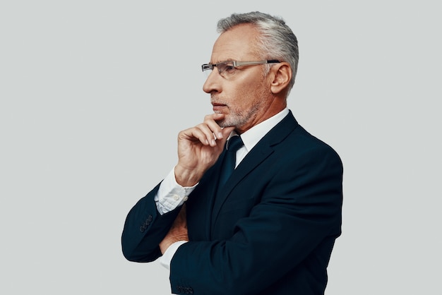 Handsome senior man in full suit looking away and keeping hand\
on chin while standing against grey background