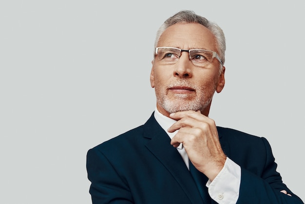 Handsome senior man in full suit looking away and keeping hand\
on chin while standing against grey background