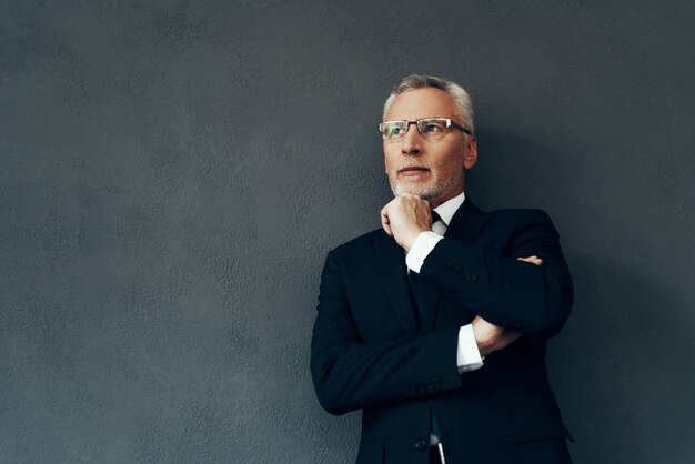 Handsome senior man in full suit looking away and keeping hand\
on chin while standing against grey background