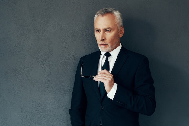 Handsome senior man in full suit thinking about something while\
standing against grey background