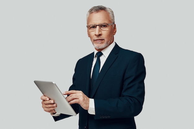 Handsome senior man in full suit using digital tablet and\
looking a camera while standing against grey background