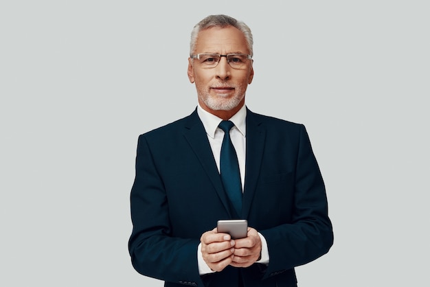 Handsome senior man in full suit using smart phone and smiling\
while standing against grey background