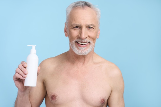 Handsome senior man showing at camera a white bottle with cosmetic beauty product isolated on blue