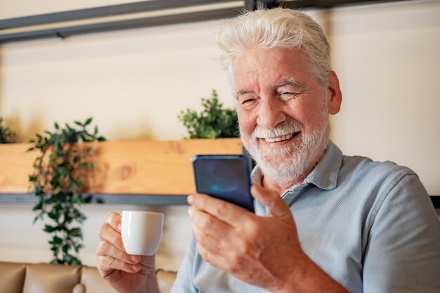 Handsome senior man sitting inside a cafeteria enjoying an\
espresso coffee cup while using mobile phone caucasian elderly\
bearded man looking at smartphone