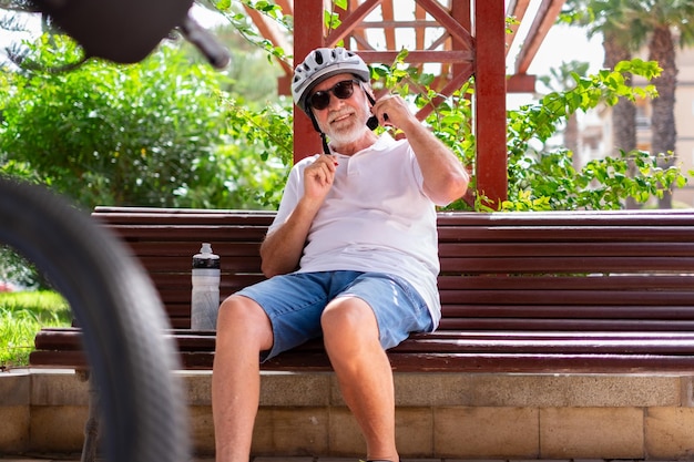 Handsome senior man with bicycle sitting in public park adjusting his helmet before to ride