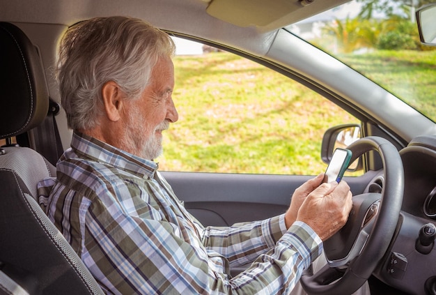 Handsome senior man with checkered shirt sitting inside his parked car using mobile phone Smiling bearded elderly grandfather with hands on steering wheel enjoying tech and social