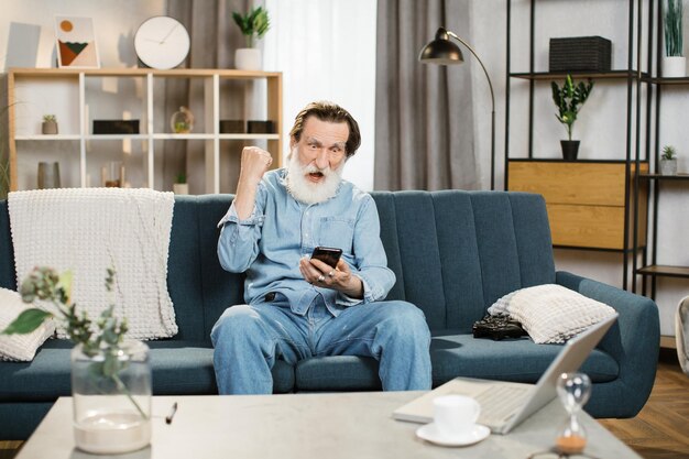 Красивый пожилой мужчина с седой бородой сидит со смартфоном на диване