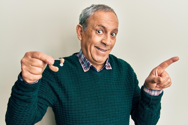 Handsome senior man with grey hair holding medical hearing aid smiling happy pointing with hand and finger to the side