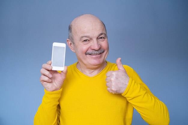 Handsome senior man in yellow sweater shows thumb up and phone screen with copyspace Studio shot on blue wall