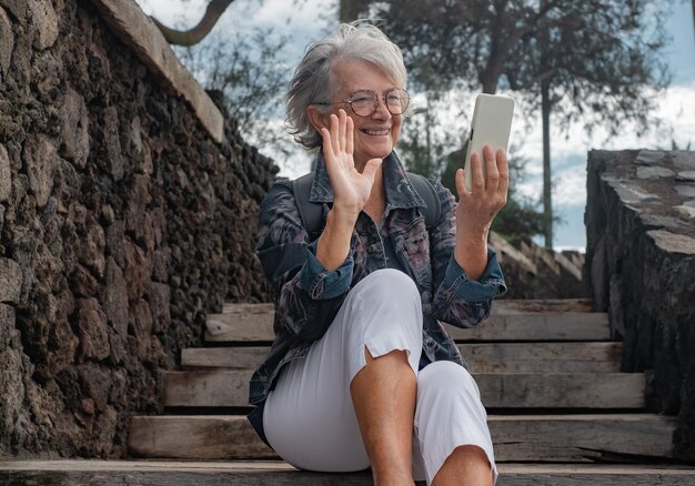 携帯電話でビデオ チャットしながら屋外の木製の階段に座っているハンサムな年配の女性 無線技術を使用して幸せな笑顔の年配の女性