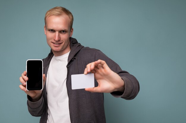 Handsome serious blonde man wearing grey sweater and white t-shirt isolated over blue background wall holding credit card and mobile phone with empty screen for cutout looking at camera.