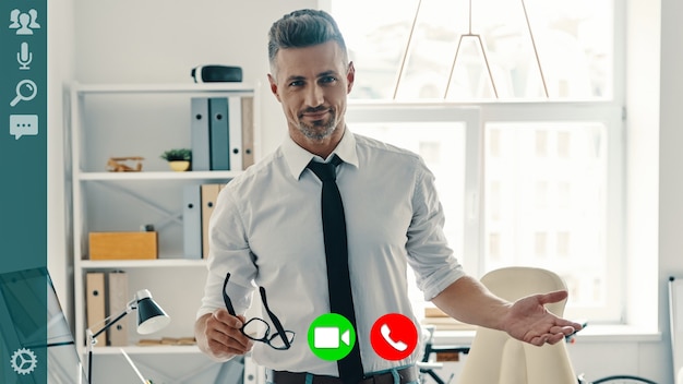 Handsome serious man in shirt and tie smiling and looking at camera while having video call