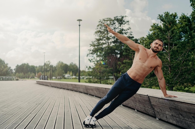 Handsome serious man stands in side plank has perfect body\
constitution poses with naked chest outdoors keeps arm raised\
trains for being healthy dressed in sport trousers and\
sneakers